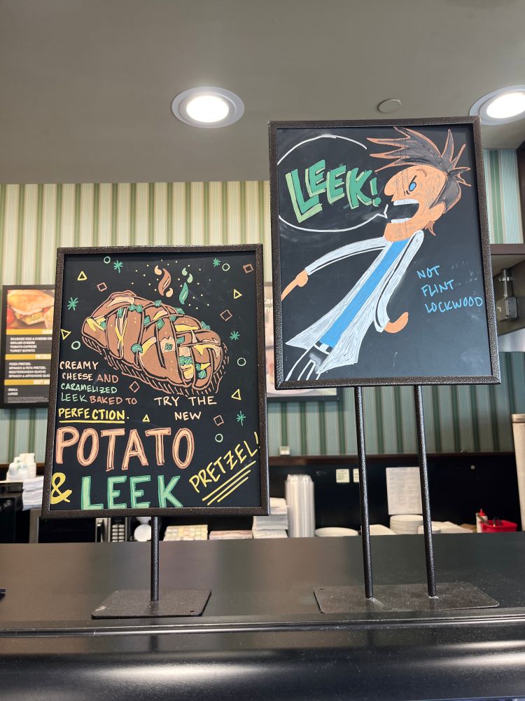 Two Starbucks signs where one sign is a drawing advertising a potato and leek pretzel. The other sign is a drawing of Flint Lockwood from the movie ‘Cloudy with a chance of meatballs’ exclaiming “LEEK!” in response to the other sign (as a reference to the movie) and a note under him that reads ‘not Flint Lockwood’ 