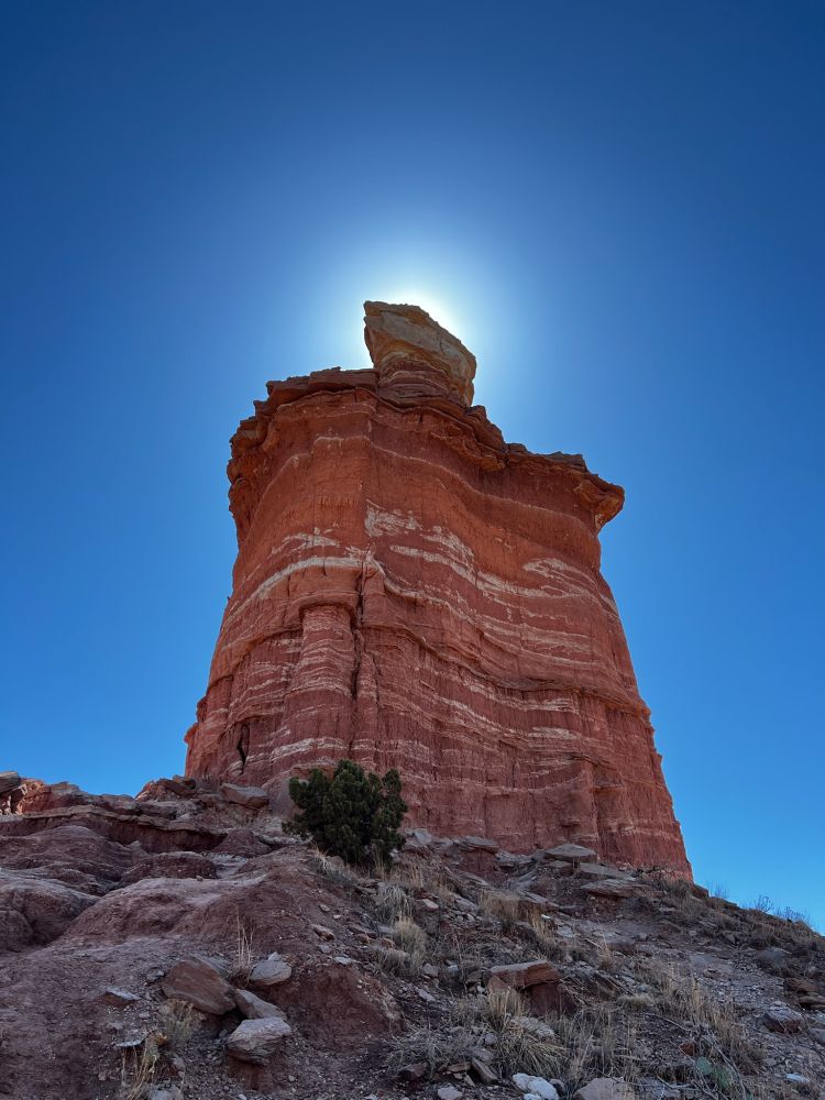 A rock tall rock formation coming out of a plateau. The sun can be seen shining just behind the top of the formation.
