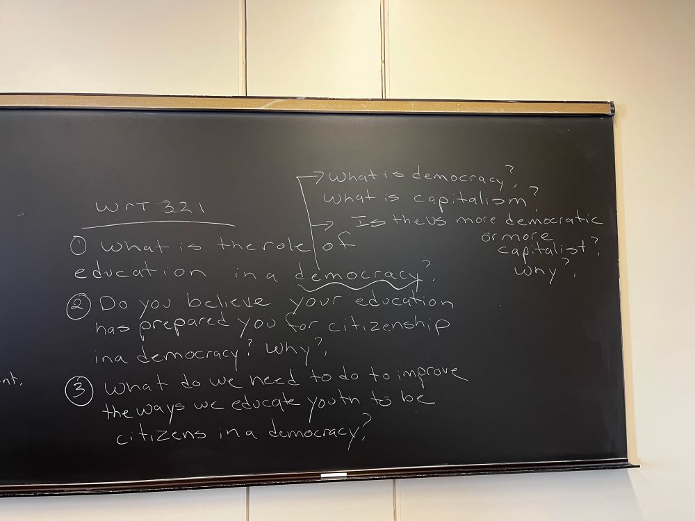 Black board with questions: what is the role of education in a democracy? Do you believe your education has prepared you for citizenship in a democracy? Why? What do we need to do to improve the ways we educate youth to be citizens in a democracy? What is democracy? What is capitalism? Is the US more democratic or capitalist? Why?