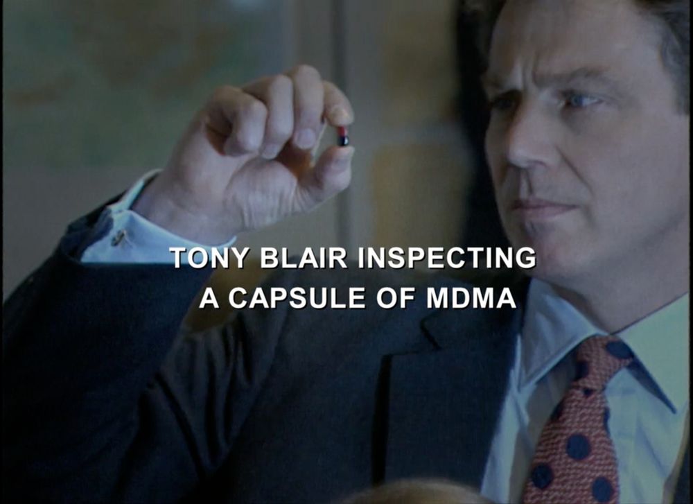 Screen shot from episode 5 of Adam Curtis's Shifty, showing former prime minister Tony Blair staring at a two-tone brown capsule he is holding between his thumb and forefinger.  Text across the image reads "TONY BLAIR INSPECTING A CAPSULE OF MDMA"