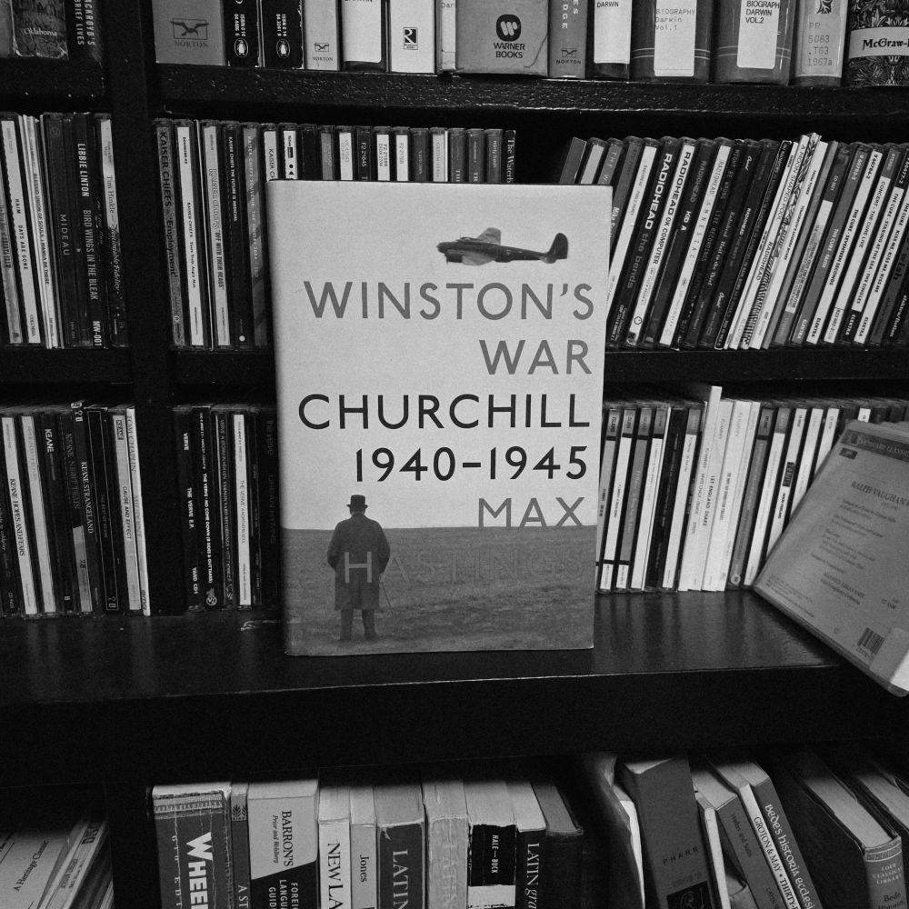 Shows the book, which features a famous photo of Churchill standing in a field watching a fighter plane fly by on its cover, standing on a shelf in front of CD spines. 