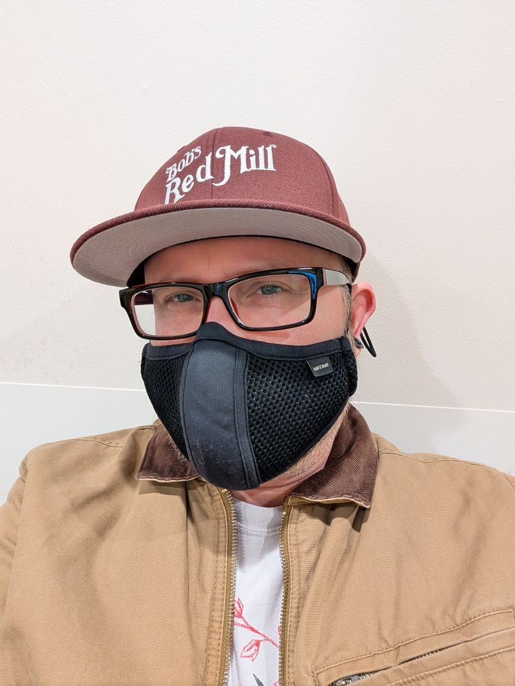 Selfie. I'm sitting against a blank wall, wearing a barncoat and Bob's Red Mill cap and facemask.