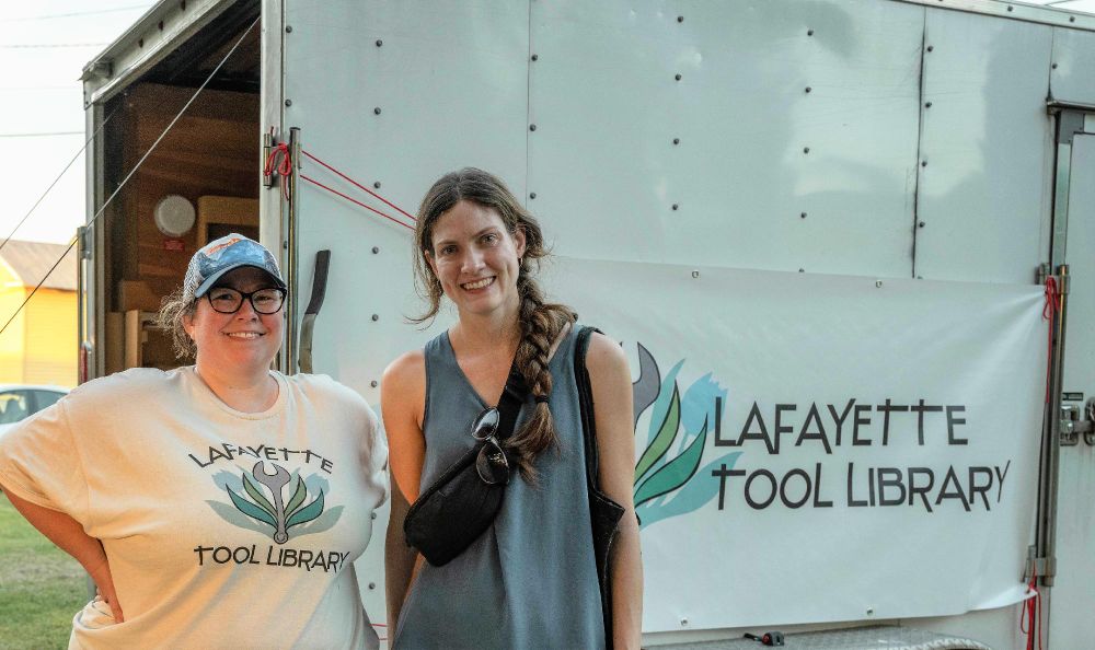 Two individuals standing in front of a trailer with a sign that reads "Lafayette Tool Library." One person is wearing a cap and a T-shirt with the library's logo, and the other is holding a bag, smiling beside them.