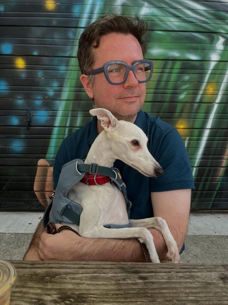 Shelby the white whippet and me, a white homosexual stare in the same direction in front of a colorful backdrop. Blues and greens abound .