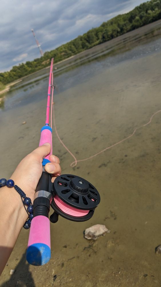 fly-fishing rod, it's pink with some blue accents on it. my arm is extended outward holding it. I'm wearing dark blue lapis lazuli beads on my wrist. the line is sitting in shallow water. this angle reveals both ends of the rod.