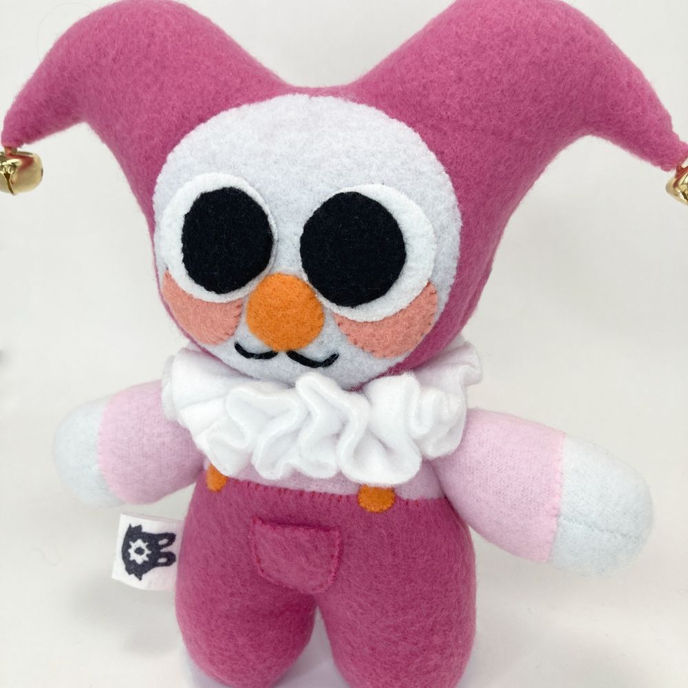 photo of a clown plushie with a pink color palette with huge eyes and bells on the end of their jester hood. a closer up photo than the previous one.