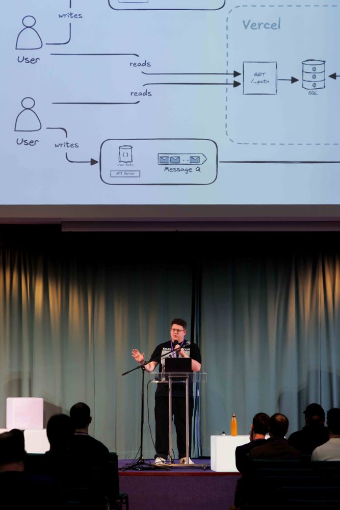Image of me standing on stage presenting a talk. I am lower center frame behind a plinth holding a microphone with one hand and gesturing with the other. Projected above me on a screen is a large diagram covering the top half of the frame