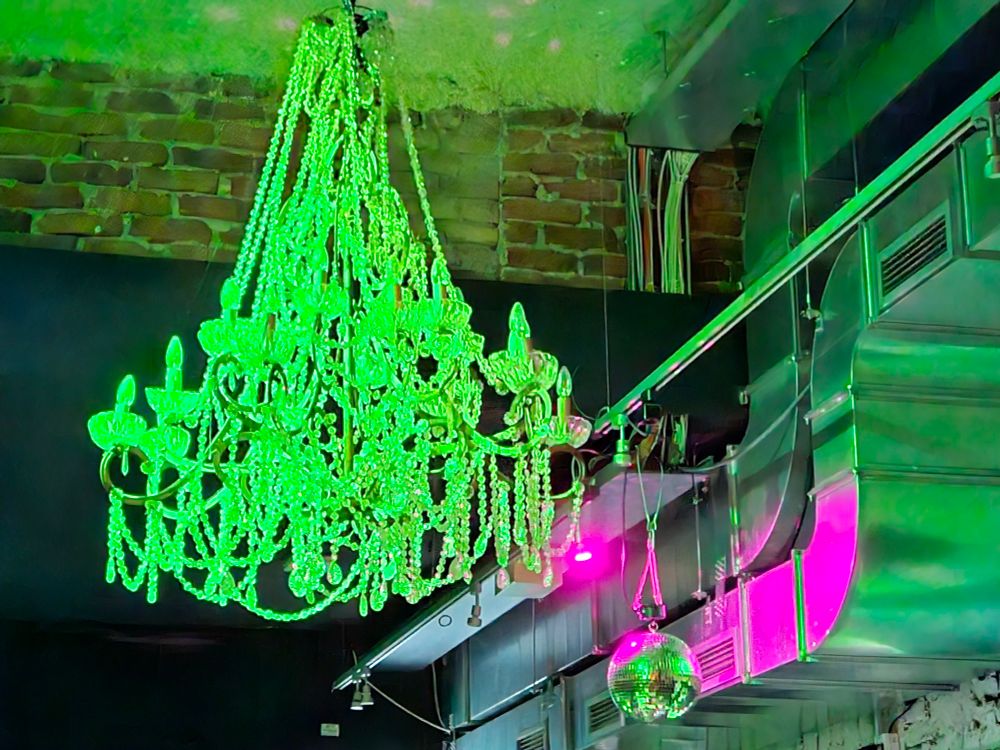 Deckenansicht eines alten Gemäuers mit metallenen Kabelkanälen und einem Kronleuchter. Discolicht - alles ist neongrün anestrahlt, punktuell ein pinker Spot. 
interior of an old building, ceiling view. metal cable trunking. a chandelier on the ceiling. neon green disco light. a smaller spotlight in the corner illuminates part of the metal in pink.