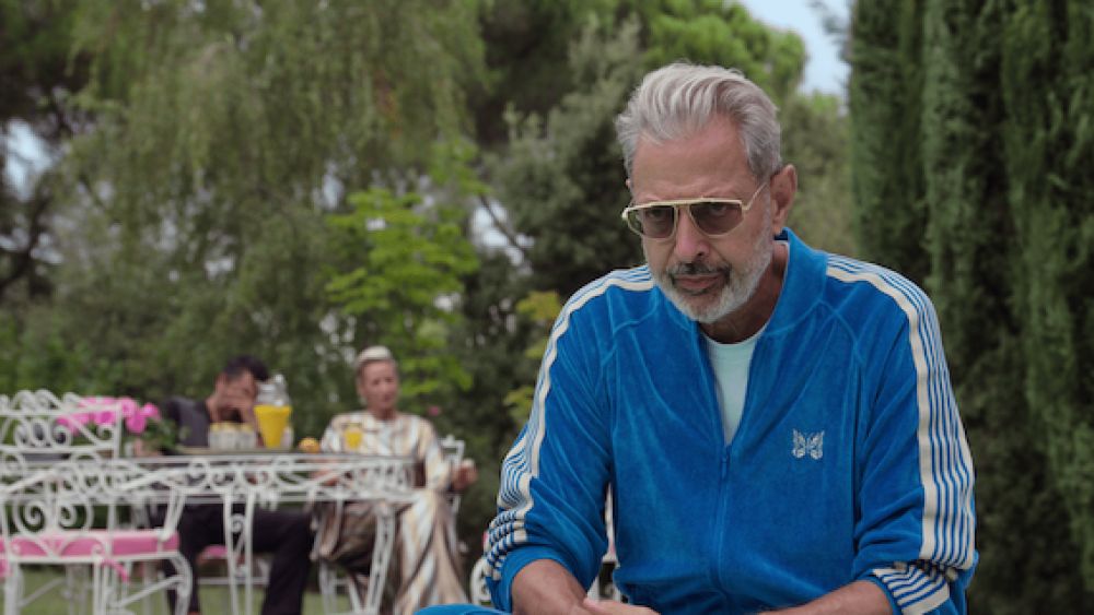 Jeff Goldblum in some TV show where he plays Zeus critically wearing a blue tracksuit from the brand Needles and some baller rich guy shades, leaning forward in seeming consternation instead of relaxing in the luxurious garden that surrounds him. There are people behind him at a table but they're too blurry for me to have any idea who they are or what they're doing 