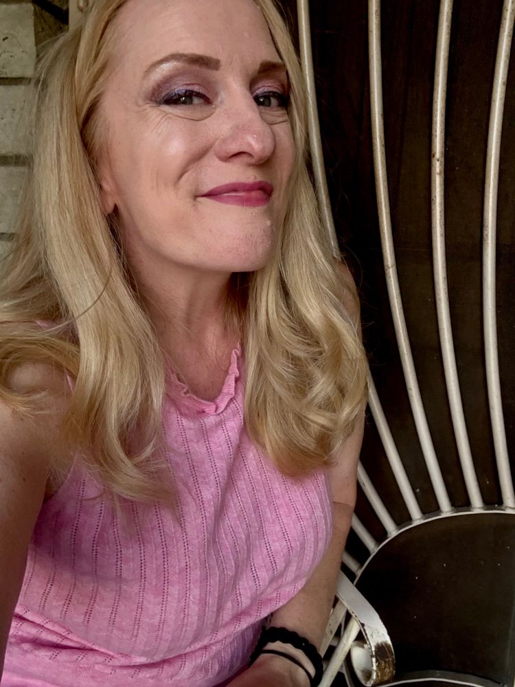 A woman with blonde hair and a pink shirt with pink and purple eye shadow and mauve lipstick is smiling in a way that looks like she already hit the edible but didn't. She wants to though 