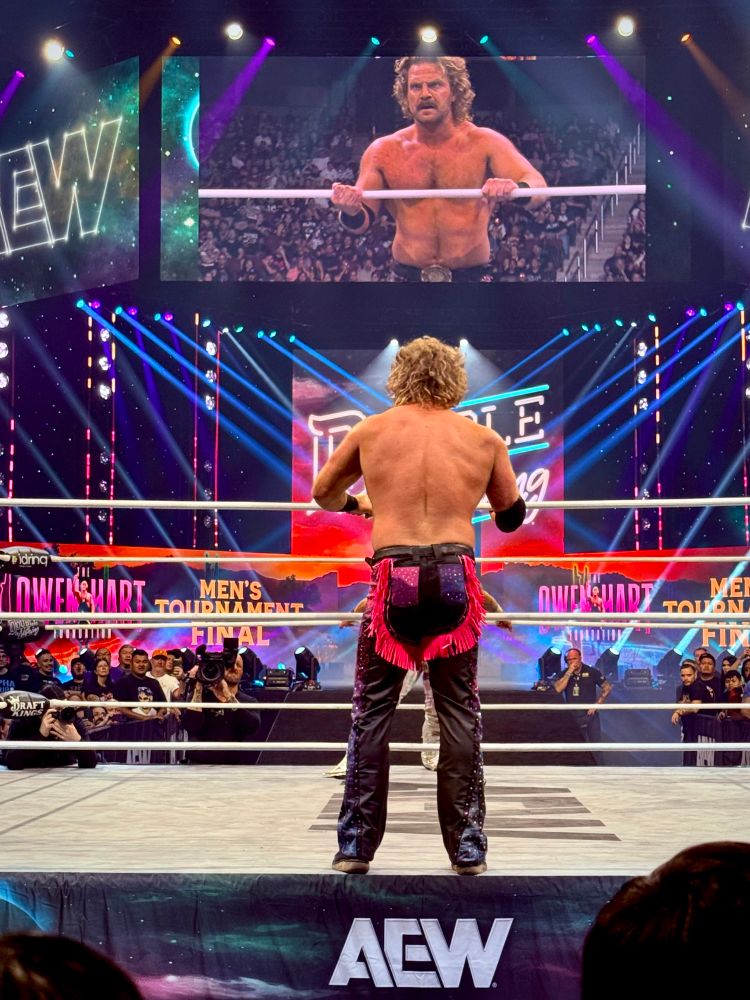 Hangman Adam Page wearing pink and black standing poised outside of the ring, waiting to hit the Buckshot Lariat onto Will Ospreay 