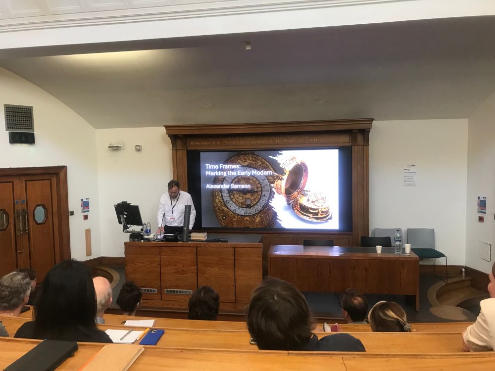Picture of a speaker in front of slides with beautiful pocket watches on it 