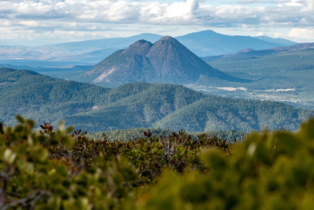 A double-humped gray summit rises behind layers of ridges cloaked in green vegetation. This is Black Butte, a small and extinct volcano on the flanks of Mt Shasta.