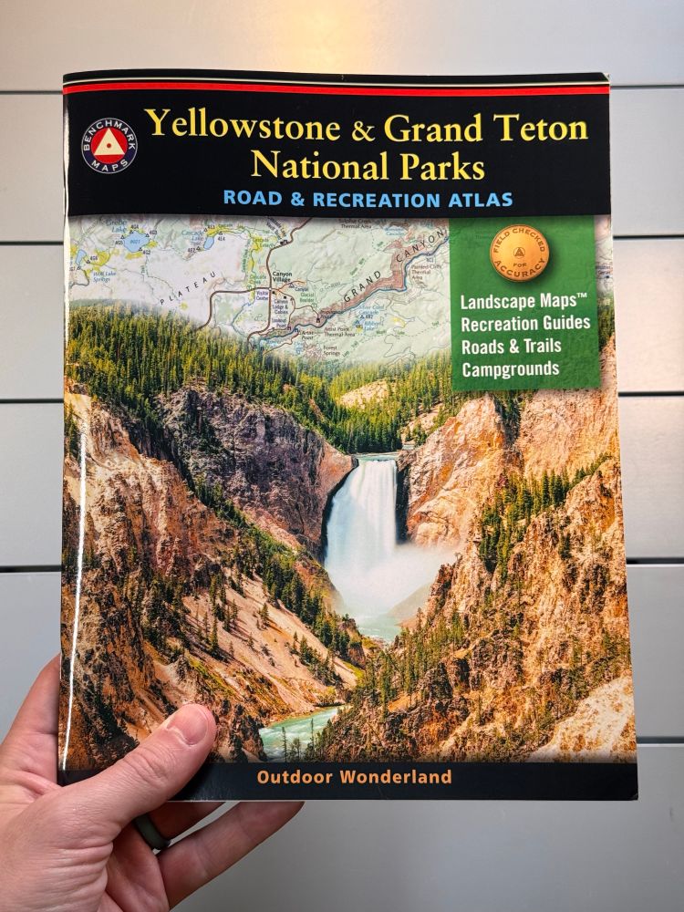 A hand holds an 8.5x11 inch book titled Yellowstone & Grand Teton National Parks Road & Recreation Atlas
