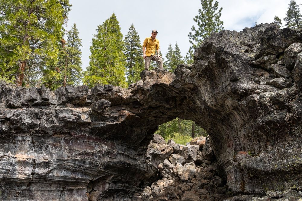 A guy in a yellow shirt (me) stands on a lava arch, leftover from a collapsed lava tube. The rock is dark gray and black. Green conifers rise over the jumble of rocks.