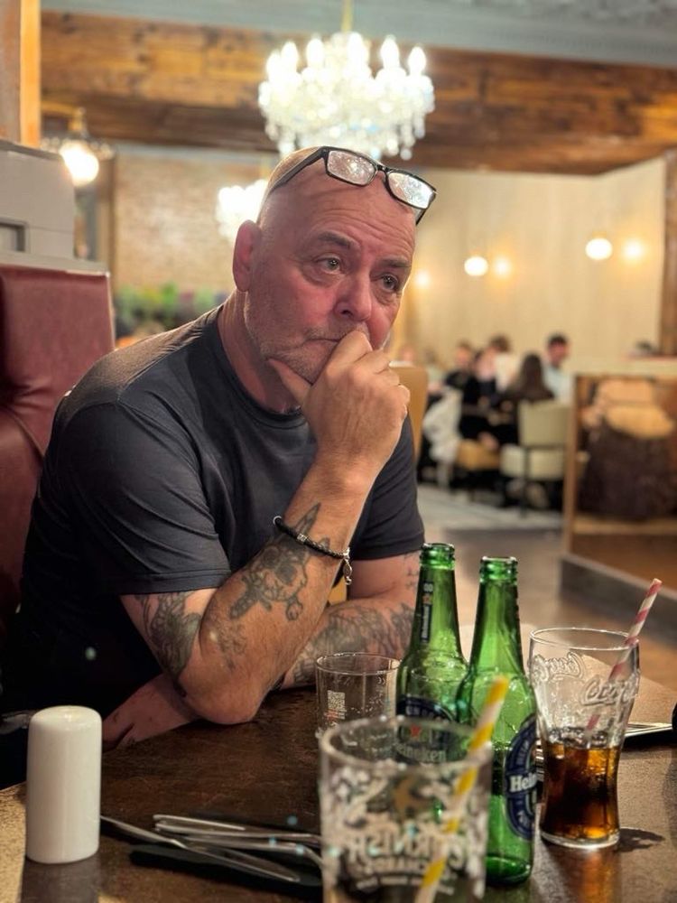 My dad, a mid fifties bald man with a Tshirt on and with his glasses resting on his forehead sitting at a restaurant table with his hand on his chin, looking deep in thought. 