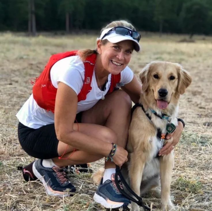 Color photograph of Heidi Carman, crouched outdoors in a grassy area, smiling warmly at the camera while embracing a happy-looking golden retriever. She is wearing a white t-shirt with a red hydration vest over it, dark shorts, and athletic shoes. The dog, a golden retriever, her therapy dog Kerith, is wearing a harness and is panting with a happy expression. The background is a natural, dry, open field with blurred trees in the distance.