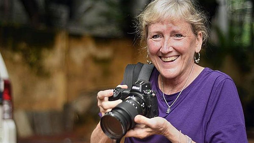 Candid image of Pulitzer Prize-winning photojournalist Carol Guzy smiling warmly at the camera while holding a Nikon professional camera with a long lens. She has short light-colored hair, wears a purple shirt and hoop earrings, and stands outdoors. The background is blurred.
