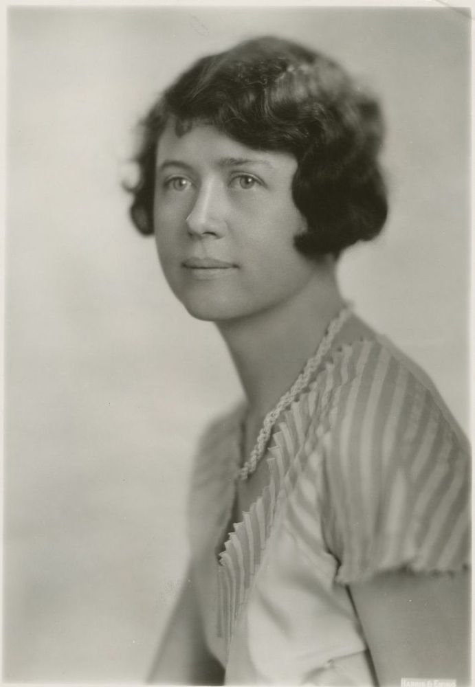 Black-and-white portrait photograph of Dr. Louella E. Cable as a young woman. She has short, dark, wavy bobbed hair styled close to her head, and is wearing a light-colored blouse with vertical pleats and a delicate necklace. She gazes calmly to the side with a gentle, confident expression against a plain studio backdrop.