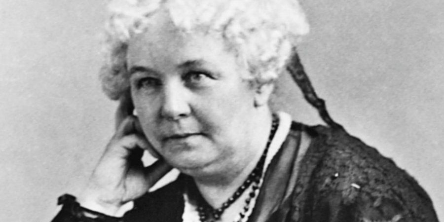 Black-and-white portrait photograph of Elizabeth Cady Stanton in her later years, circa 1890s. She gazes thoughtfully to the side with her chin resting gently on her right hand, wearing a dark Victorian dress with a high lace collar and long beaded necklace. Her white hair is styled in soft curls framing her face, against a plain studio backdrop. The image conveys quiet strength and determination, characteristic of the pioneering women’s rights activist and suffragist leader.