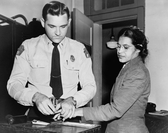 Black-and-white photograph from December 1, 1955: Rosa Parks, standing and wearing glasses and a coat, calmly has her fingerprints taken by a white police officer in Montgomery, Alabama, shortly after her arrest for refusing to give up her bus seat to a white passenger—an act that sparked the Montgomery Bus Boycott and became a defining moment of the Civil Rights Movement.