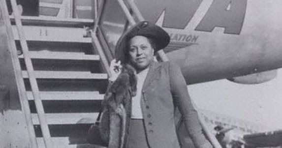 Black-and-white photo of Edith Sampson at the bottom of the stairs about to board a plane. She is wearing a suit and has on a very handsome hat.