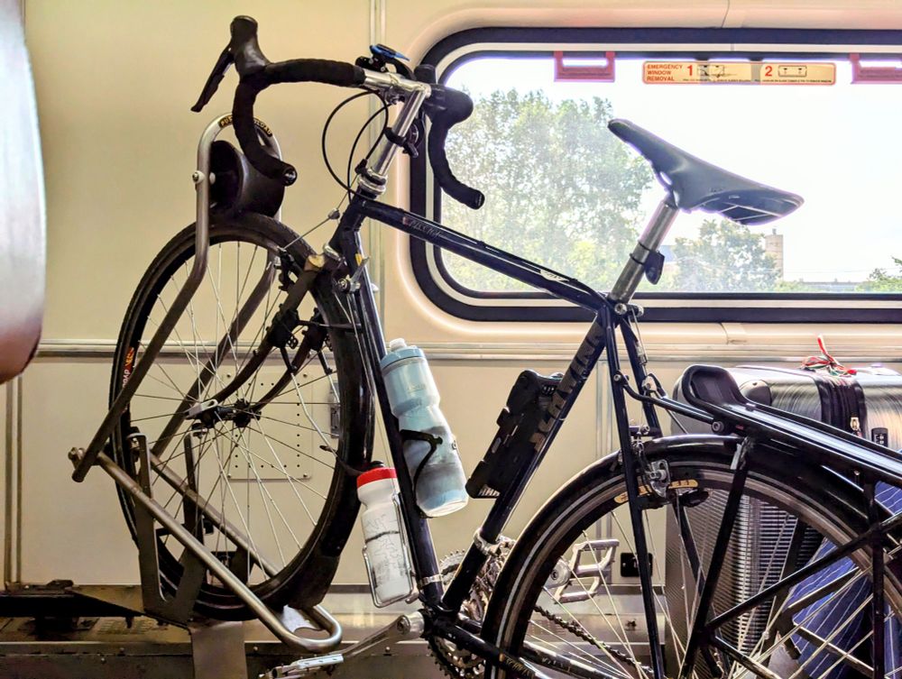 Photo of a bicycle in a rack inside a train with part of the trail window visible and a tree outside.