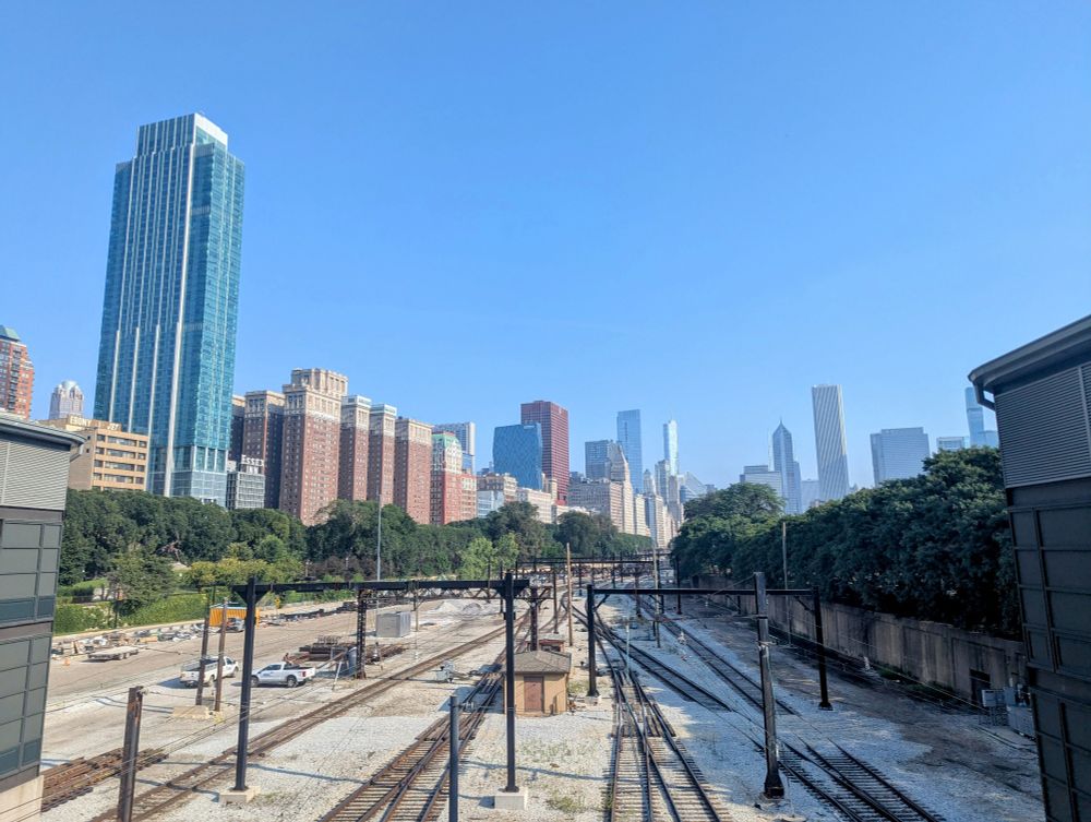 Photo looking out over railroad tracks leading towards the tall buildings of the Chicago skyline under a blue sky.