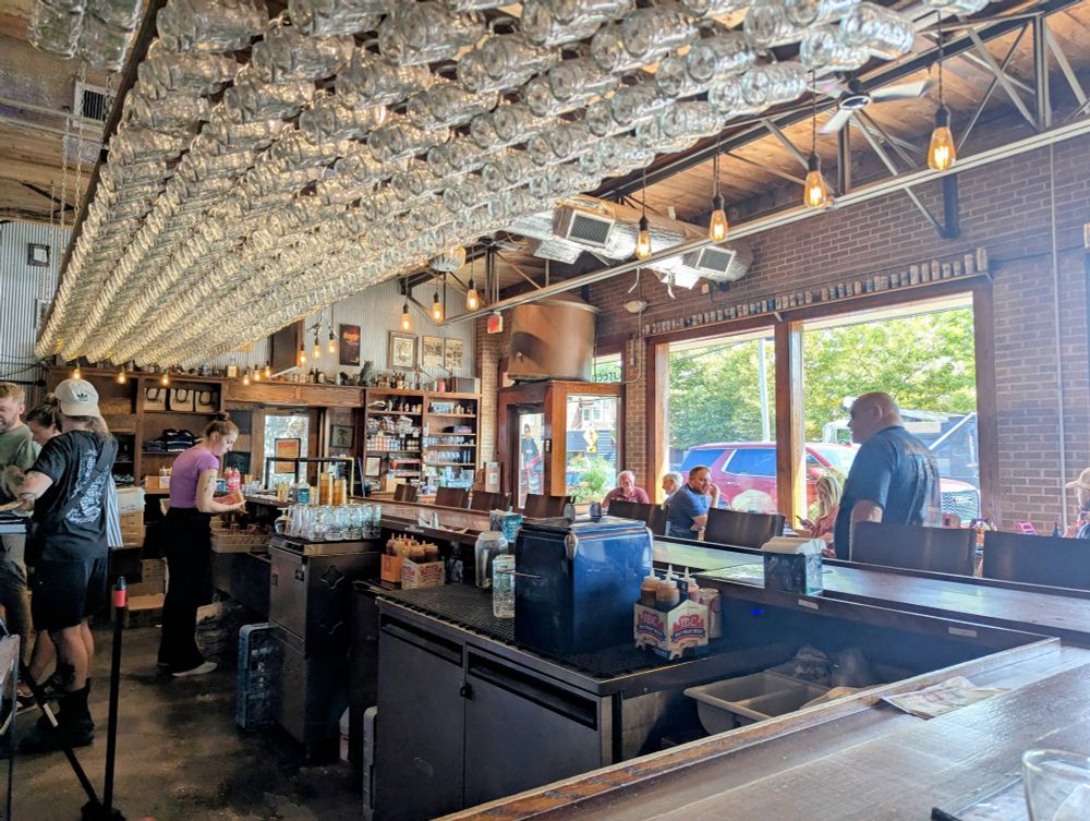 Photo taken across the corner of a bar inside a brewery in Sawyer, Michigan called Greenbush. Overhead is a rack full of glass beer mugs.