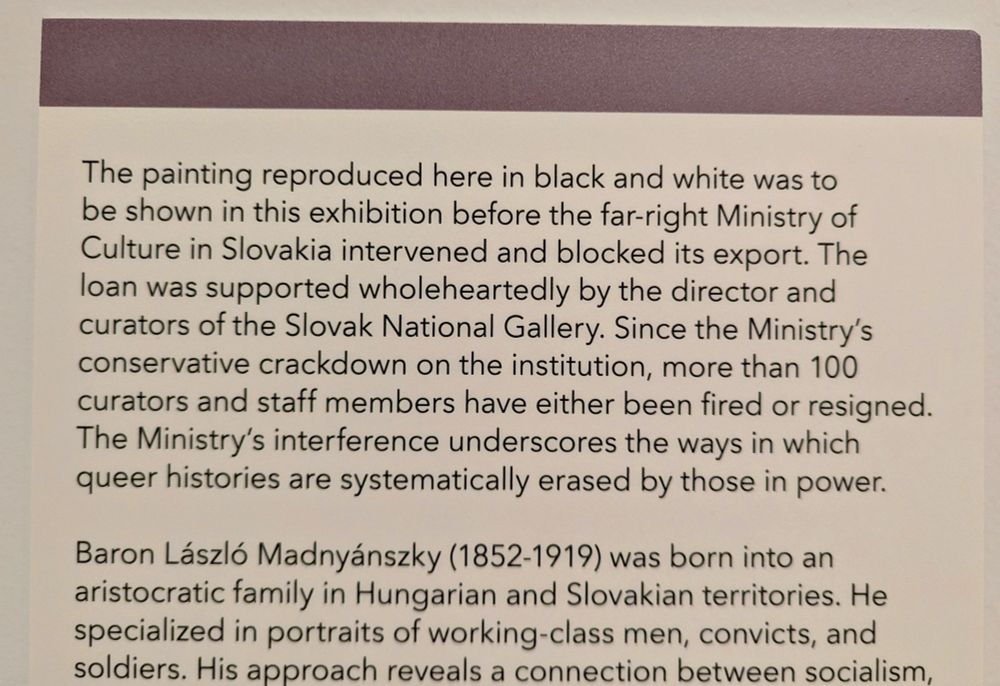 Photo of text on a wall mounted sign at an exhibit that reads: the painting reproduced here in black and white was to be shown in this exhibition before the far right ministry of culture in Slovakia intervened and blocked its export. The loan was supported wholeheartedly by the director and curators of the Slovak national gallery. Since the ministry's conservative crackdown on the institution, more than 100 curators and staff members have either been fired or resigned. The ministry's interference underscores the ways in which queer histories are systematically erased by those in power.