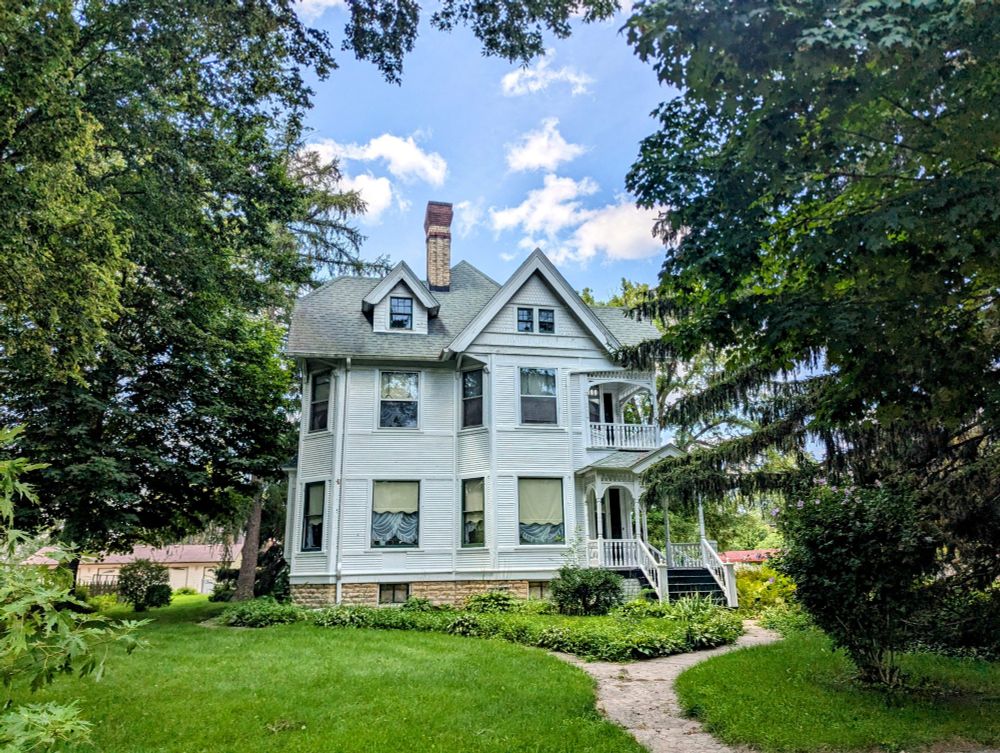 Photo of a three-story Victorian house on a lush lot painted mostly pale off-white.