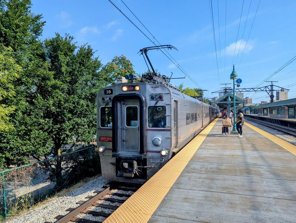 Photo of the front of an electric train pulling into a high-level station platform under a mostly clear blue sky.