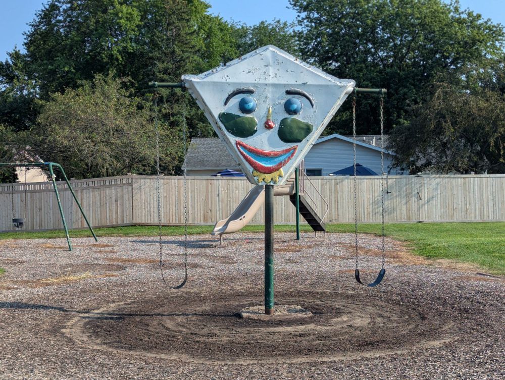 Photo of a white diamond shaped object perched on a pole with a colorful clown face painted on it and swings dangling off of arms protruding to its left and right on a playground.
