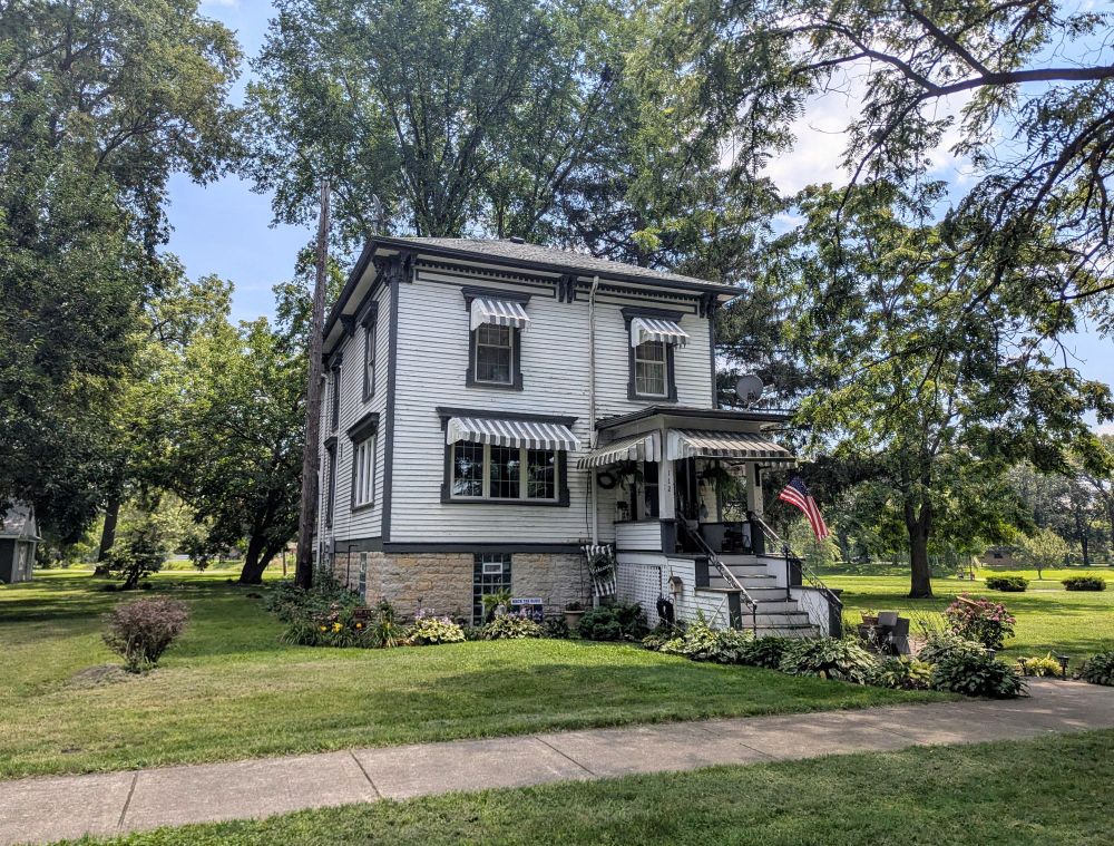 Photo of a two-story italianate house with dark trim and light wood siding and awnings over the windows on the front.