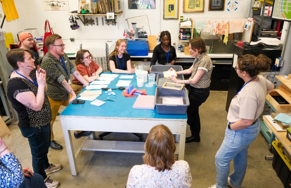A group of participants circles a blue table as they watch an instructor demonstrate papermaking.