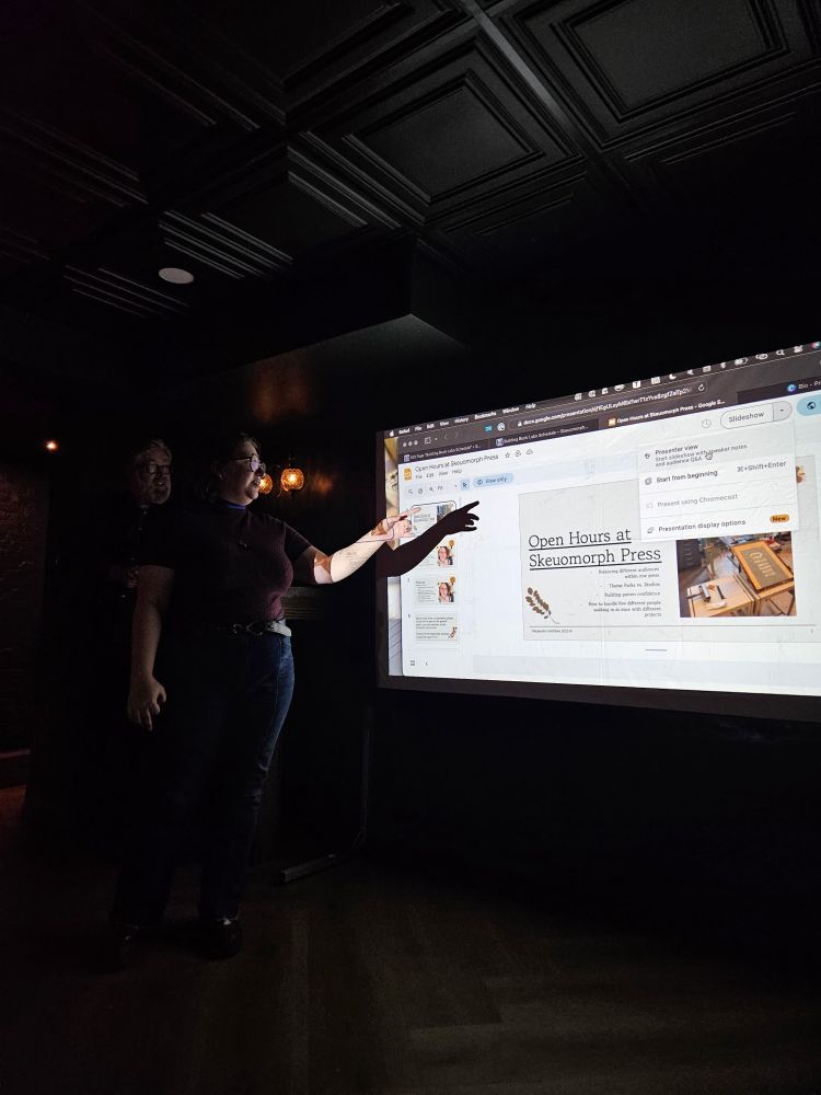 A woman with glasses mainly in shadow gestures to a power point in a dark speakeasy. The title slide reads “Open Hours at Skeuomorph Press”.