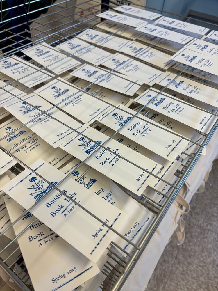 A drying rack full of white bookmarks reading in blue ink “Building Book Labs, a book is… Spring 2025”. There is an image on the bookmarks of a flower growing out of a book.