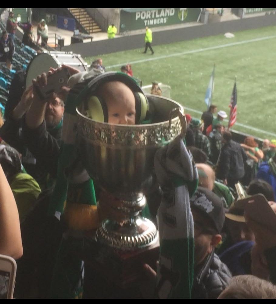 A baby safely (?) tucked into a silver trophy cup, surrounded by sports fans. 