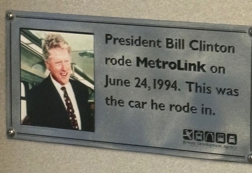 A picture of a silver-colored plaque on a white train car wall that reads "President Bill Clinton rode MetroLink on June 24, 1994. This was the car he rode in." To the left of the text is a picture of Bill Clinton standing in the very same MetroLink train car.