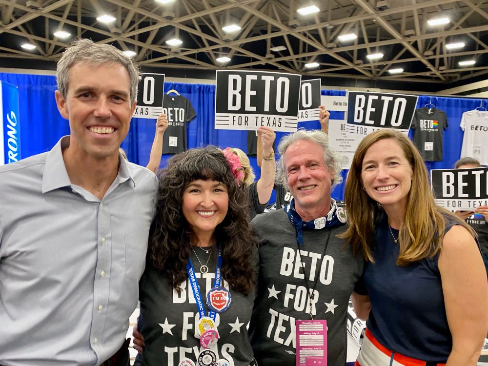 Tall gray haired man in blue button down shirt, woman with long dark curly hair and black & white tee, gray haired man with black & white tee, woman with long blonde hair and blue top, all smiling in front of a blue curtain and Beto For Texas signs.
