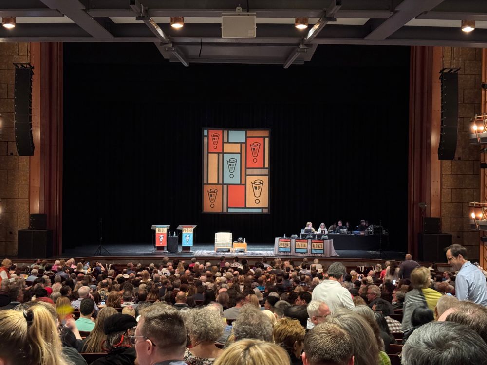 An audience of people in a theatre, about to watch a show.  Two lecterns with signs with the hosts’ names, a comfy chair and side table, and a panelist table with 3 name placards hanging off the front of the table.  In the back, a large hanging sign with colored blocks with wood tone creating a mosaic, with some of them with the show’s logo, a rolled up newspaper and a circle creating an exclamation mark !.  To the right rear, the control table with audio equipment.