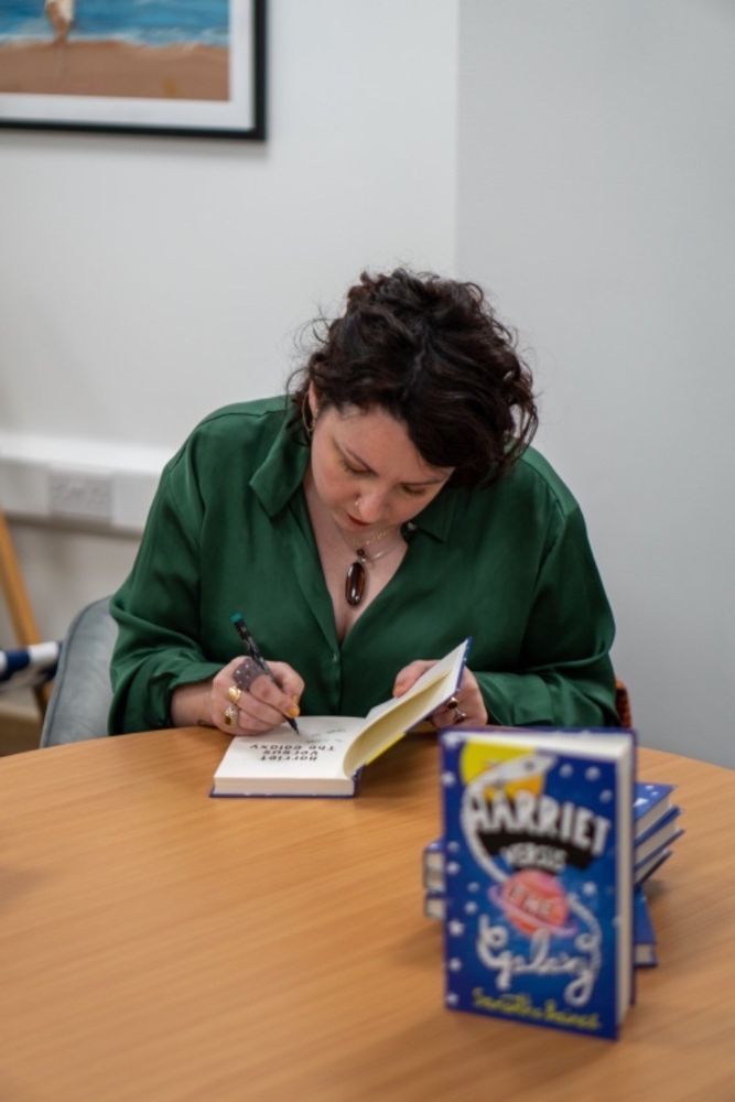 Samantha signs a copy of her book Harriet’s versus the Galaxy. She is white with black curly hair and her face is leaning over her book signing. She is wearing a silky green shirt and sitting at a wooden table with a pile of her books in front of her