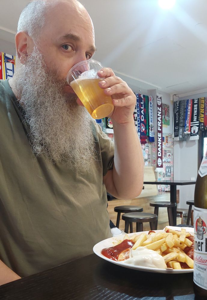 En skäggig man äter currywurst o dricker pilsner. 