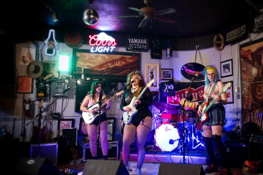 Three femmes up front and a masc drummer, Bad Buddy hits the stage at Tracks Pub in Olds, Alberta. The small town, dive bar vibe is apparent. 