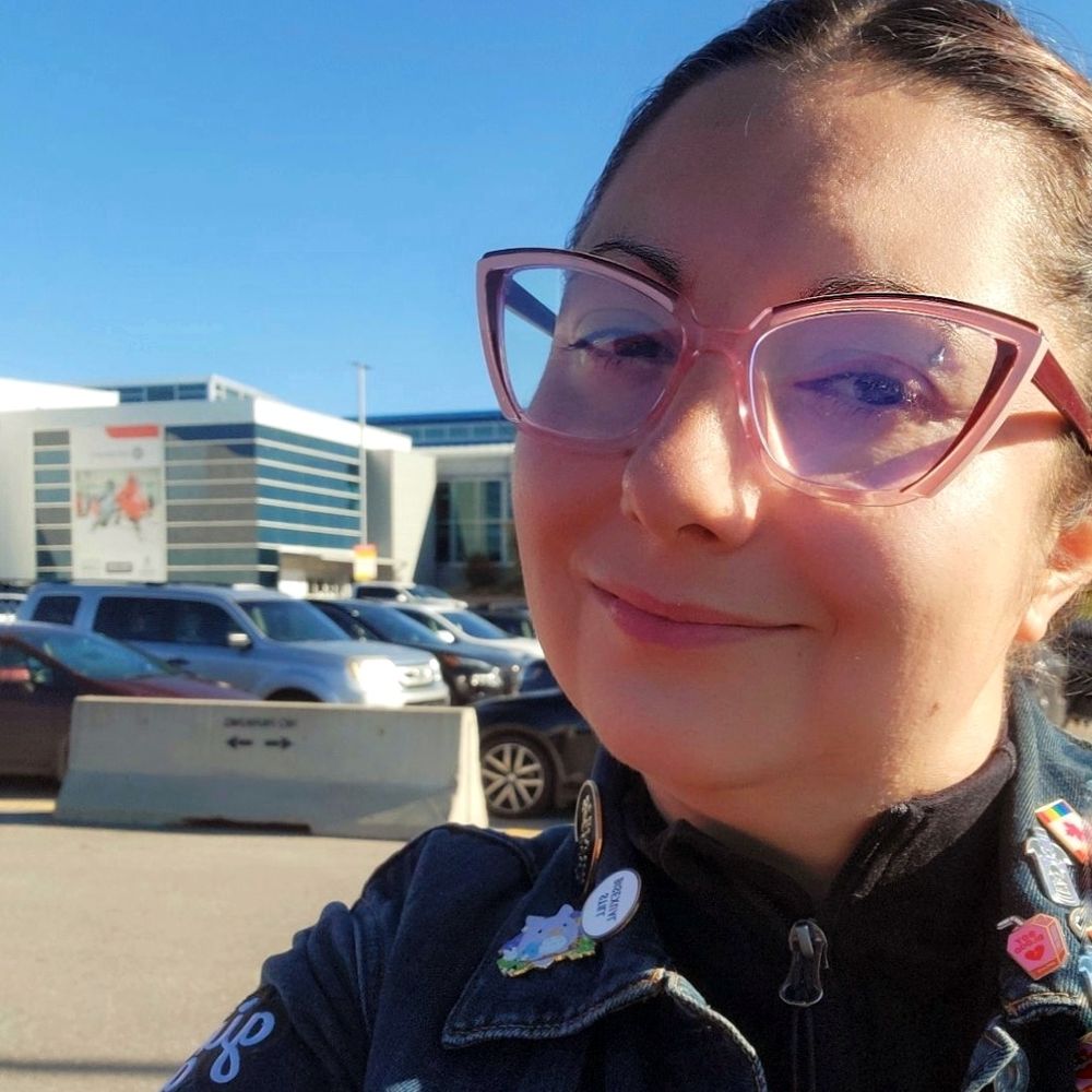 A femme human smiles in an arena parking lot. They are wearing pink and purple glasses, and a jean jacket with lots of pins and badges.