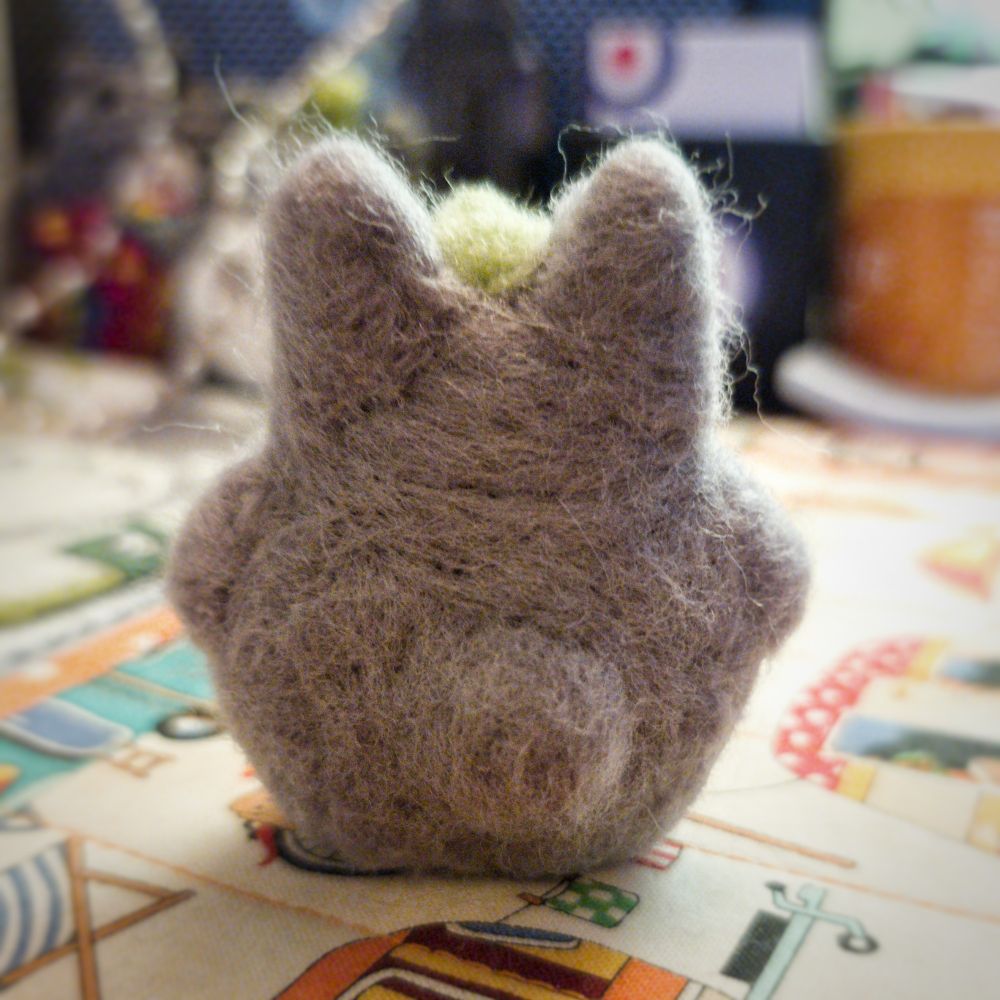 A small, felted grey Totoro wearing a green leaf as a hat. Back view.