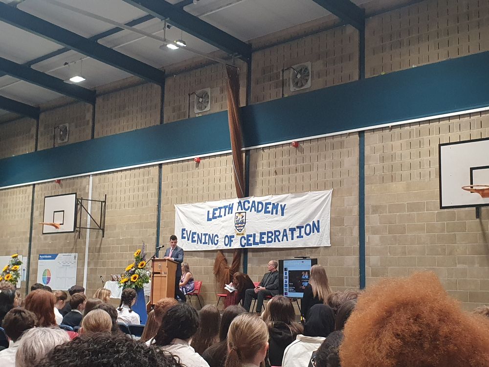 A packed gym hall, with a man speaking at a podium in front of a banner which reads: 'Leith Academy, Evening of Celebration'