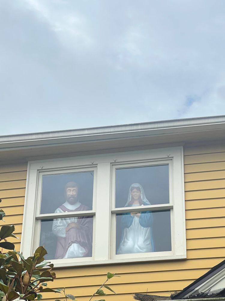 Blow molds of Joseph and Mary, each looking towards the street from the separate windows of a yellow clapboard house 