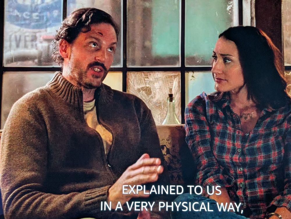 Monroe and Rosalee are sitting on a couch.  Monroe is saying "explained to us in a very physical way."  He's wearing a brown knit zip up sweater.