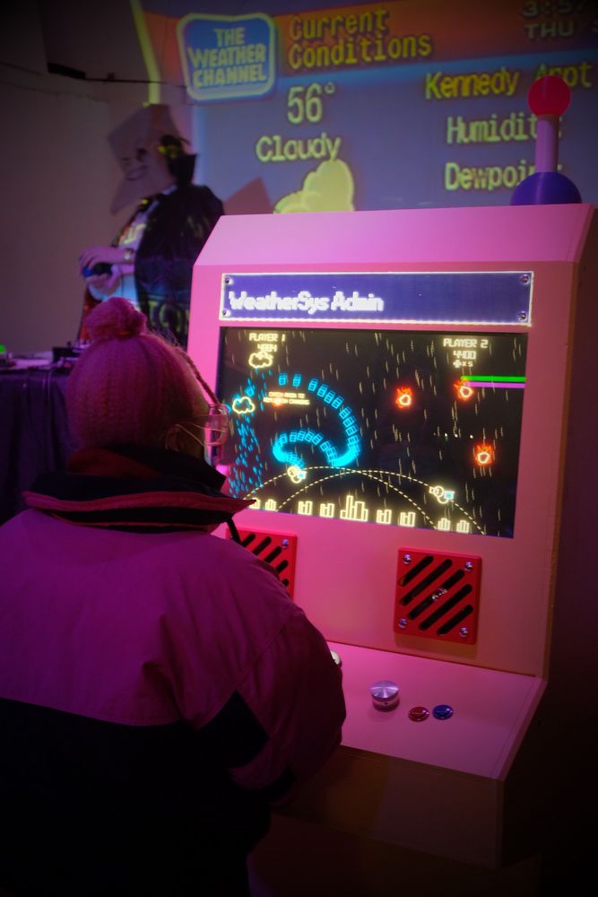 A person plays the WeatherSys Admin cab during the launch party - a DJ with a paper bag over their head plays on the stage behind the cabinet.